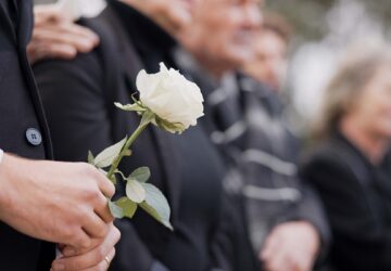 Hands, rose and a person at a funeral in a cemetery in grief while mourning loss at a memorial service. Death, flower and an adult in a suit at a graveyard in a crowd for an outdoor burial closeup
