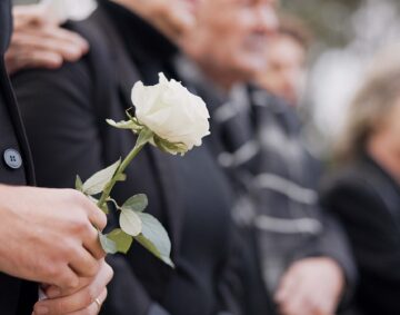 Hands, rose and a person at a funeral in a cemetery in grief while mourning loss at a memorial service. Death, flower and an adult in a suit at a graveyard in a crowd for an outdoor burial closeup