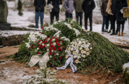 Valmiera, Latvia - January 6, 2022 - A fresh grave covered with evergreen branches and floral wreaths, surrounded by mourners in a snowy cemetery during a winter funeral.