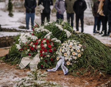 Valmiera, Latvia - January 6, 2022 - A fresh grave covered with evergreen branches and floral wreaths, surrounded by mourners in a snowy cemetery during a winter funeral.