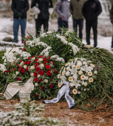 Valmiera, Latvia - January 6, 2022 - A fresh grave covered with evergreen branches and floral wreaths, surrounded by mourners in a snowy cemetery during a winter funeral.